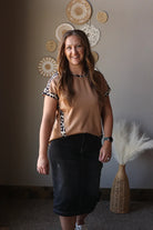 Woman standing in a room with decorative wall art and a vase with pampas grass. wearing a brown leopard short sleeve top and black denim skirt