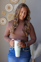 A woman with long wavy brown hair wearing a brown long-sleeve mock neck top and blue jean skirt, holding a mint green insulated travel tumbler with a dark lid. She is standing indoors against a gray wall decorated with circular woven wall art.