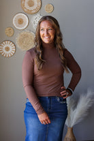 A smiling woman with long wavy brown hair stands indoors wearing a fitted long-sleeve brown mock-neck top and blue jean skirt. She is posed against a gray wall decorated with woven circular wall art, with a vase of pampas grass visible at the lower right.