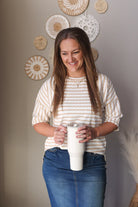 Woman holding a white tumbler, wearing a beige white striped half sleeve top for casual spring office outfits, against a neutral wall with decorative plates.