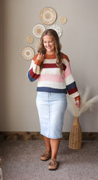 A woman stands indoors wearing a chunky knit striped sweater with horizontal bands of rust orange, cream, light pink, navy, and burgundy, paired with a light blue pencil skirt and brown lace-up shoes. The background shows a neutral beige wall with decorative woven wall art and a woven vase with dried pampas grass.