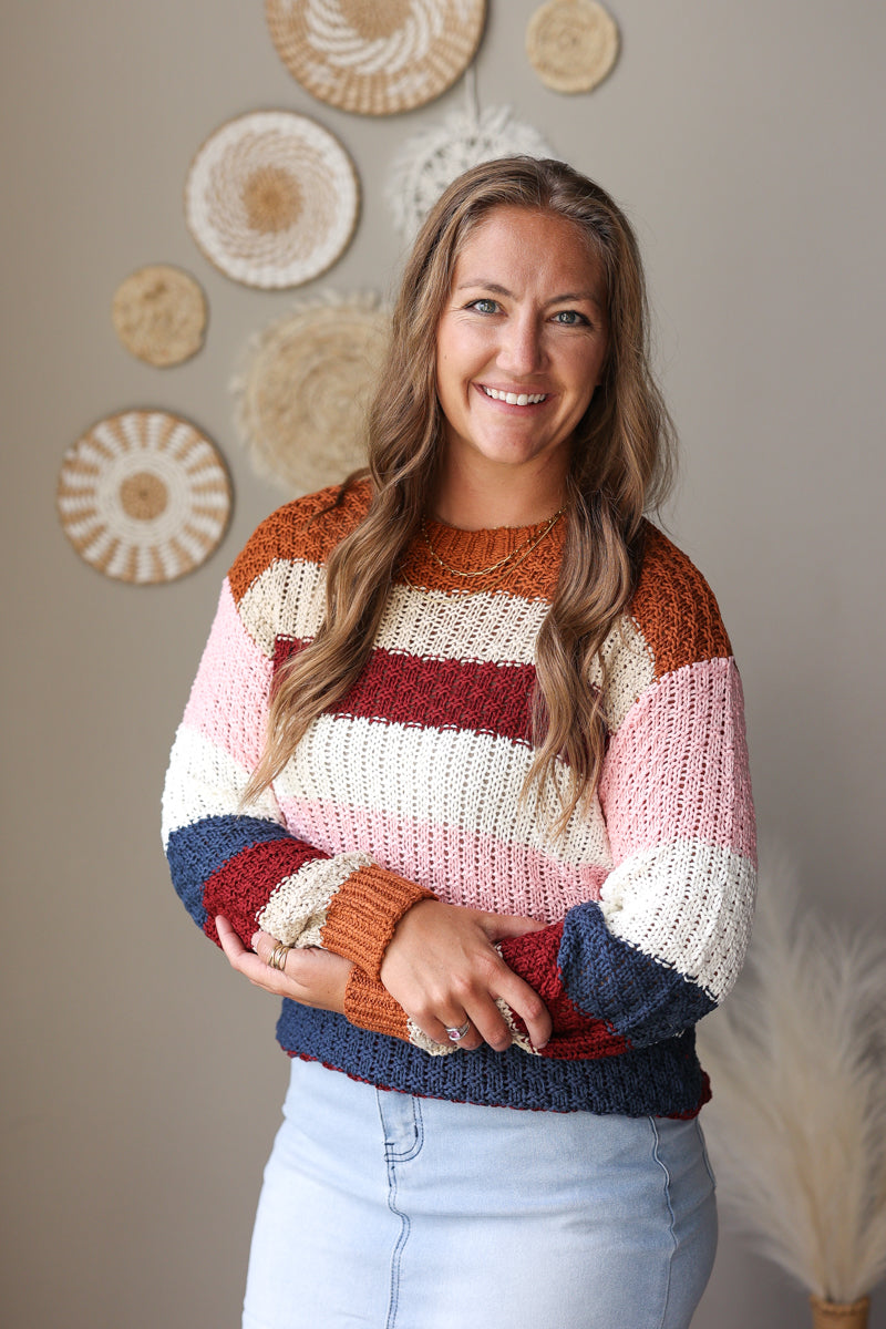 A smiling woman with long light brown hair wears a chunky knit striped sweater with horizontal bands of rust orange, cream, pink, burgundy, and navy blue, paired with light blue jean skirt. She is standing against a warm gray wall decorated with circular woven wall art.