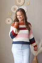 A woman wearing a chunky knit striped sweater with horizontal bands of cream, rust orange, burgundy, light pink, and navy blue, paired with light-wash blue jean skirt. She is standing in a room with a beige wall decorated with circular woven wall art and a vase with dried grass.