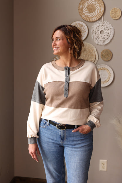 Woman wearing a color-blocked sweater and jeans standing in a room with decorative wall hangings. The Neutral Colorblock Waffle Henley Top offers cozy warmth with its soft thermal waffle fabric, perfect for effortless layering or standalone wear.
