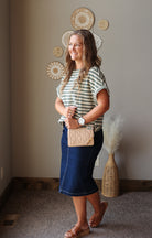 A woman posing in a home interior wearing a green and white striped t-shirt, a dark blue denim midi skirt, and brown leather sandals, while holding a beige quilted crossbody bag.