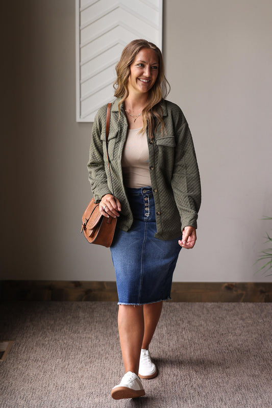 Woman wearing a green jacket, white top, blue denim skirt, and white shoes indoors.