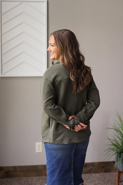 Woman wearing a green sweater and blue jeans standing in a room with a plant and wall art.