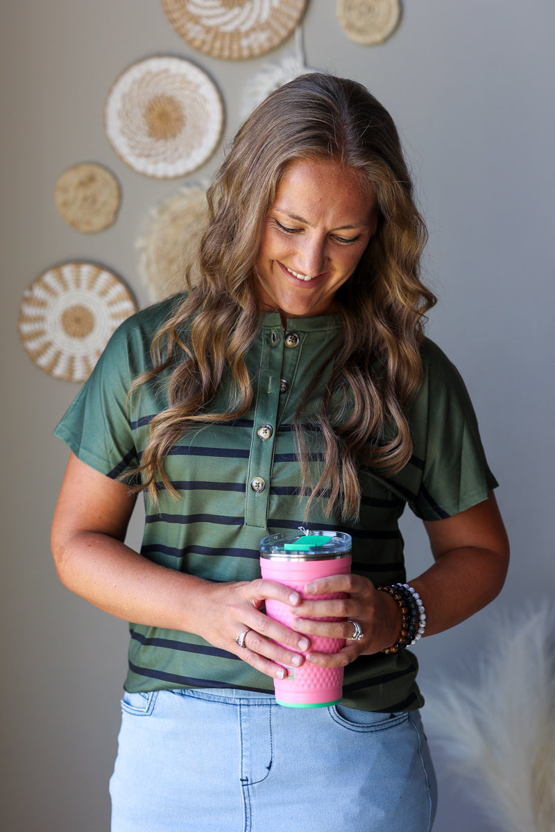 A woman with long wavy brown hair is shown from the waist up, wearing a green short-sleeve button-up shirt with thin dark horizontal stripes and light blue jean skirt. She is holding a pink reusable travel tumbler with a clear lid and a green straw.
