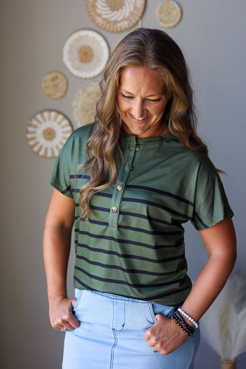 A woman wearing a green short-sleeve henley shirt with dark horizontal stripes and light blue jean skirt, standing against a gray wall with decorative circular wall art.