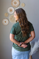A woman with long brown hair is shown from the back wearing an olive green short-sleeve t-shirt and light blue jean skirt, standing against a gray wall decorated with circular woven wall art.