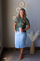 A woman stands indoors wearing a green and black striped short-sleeve top with a light blue denim skirt, holding a pink cup. The room has a gray wall with decorative woven wall art, a carpeted floor, and a woven vase with dried pampas grass.