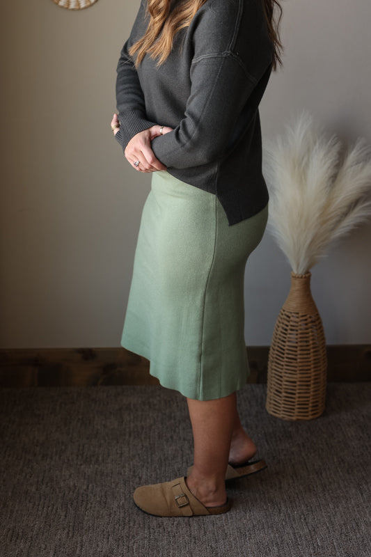 Person wearing a green skirt and dark sweater standing in a room with a textured wall and decorative vase.