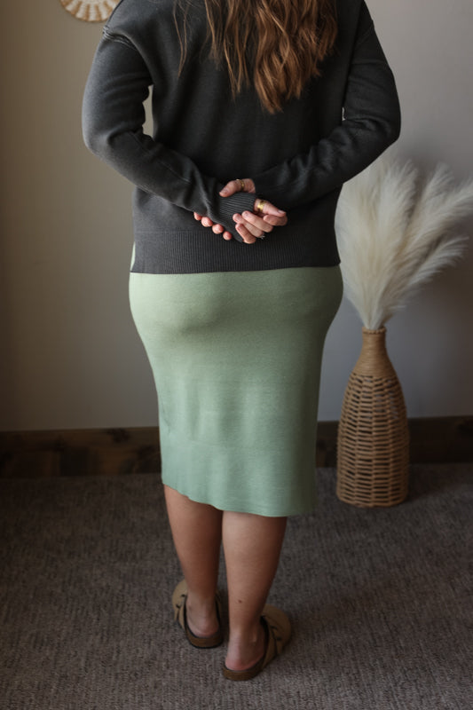 Person wearing a dark sweater and green skirt standing in a room with a textured wall and decorative vase.