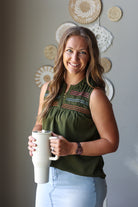 A smiling woman with long light brown hair stands indoors holding a tall white travel mug. She wears a sleeveless olive green top with colorful embroidered detailing across the chest and light blue jean skirt.