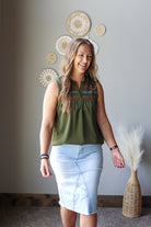 A woman modeling a sleeveless olive green blouse with colorful embroidered detailing around the neckline, paired with a light blue denim pencil skirt, standing in a room with boho wall decor.