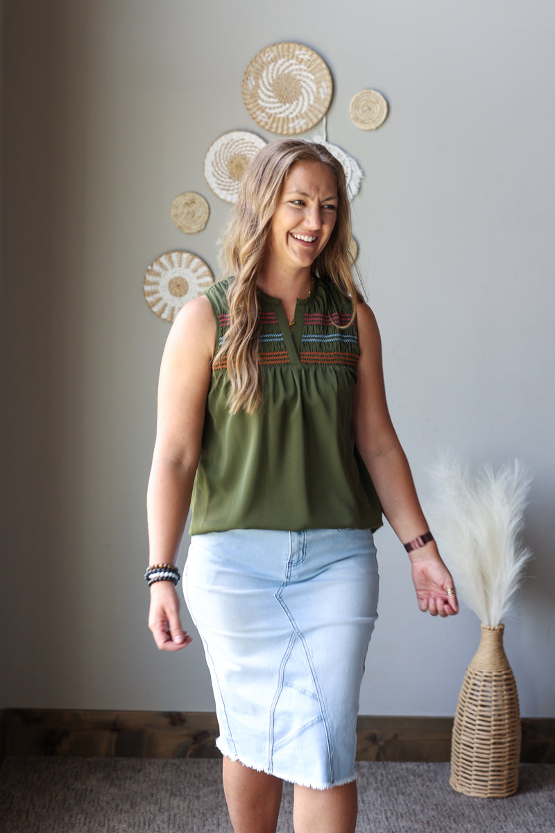 A woman modeling a sleeveless olive green blouse with colorful embroidered detailing around the neckline, paired with a light blue denim pencil skirt, standing in a room with boho wall decor.