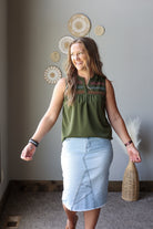 A woman modeling a sleeveless olive green top with colorful embroidered detailing at the neckline and a light blue denim skirt, standing in a room with boho-style wall decor.