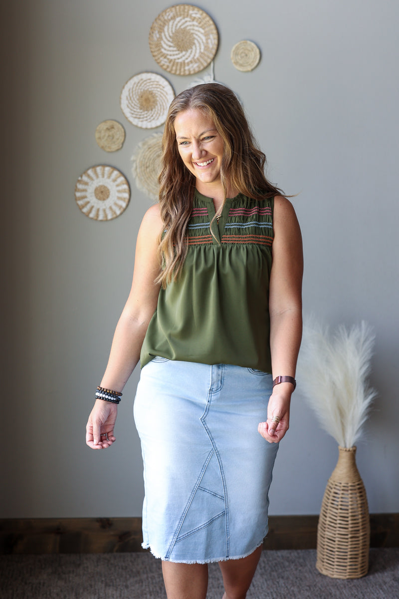 A woman modeling a sleeveless olive green top with colorful embroidered trim and a light blue denim skirt, standing in a room with boho wall decor.