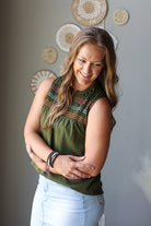 A woman with long light brown hair is wearing an olive green sleeveless top with colorful embroidered details across the chest, paired with light blue jean skirt. She is standing against a gray wall decorated with circular woven wall art.