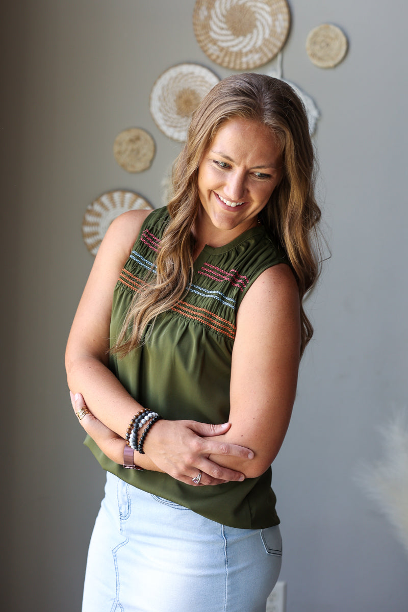A woman with long light brown hair is wearing an olive green sleeveless top with colorful embroidered details across the chest, paired with light blue jean skirt. She is standing against a gray wall decorated with circular woven wall art.