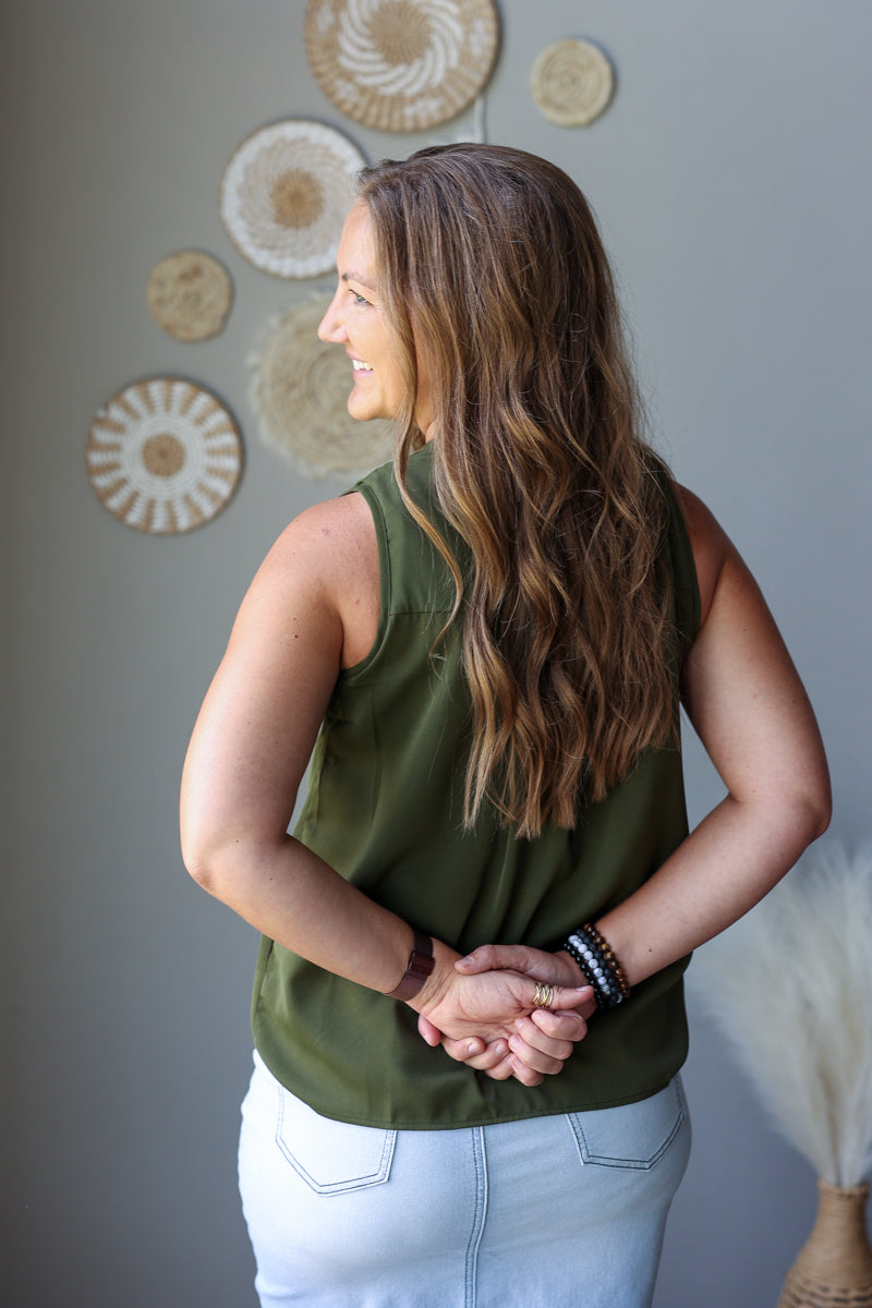 A woman with long wavy brown hair is shown from the back wearing an olive green sleeveless top and light wash denim skirt, standing against a gray wall with decorative woven wall art.