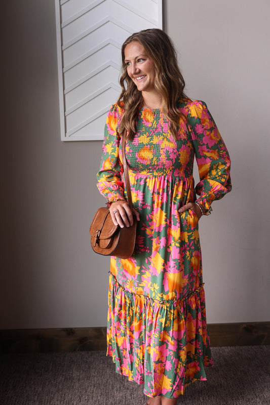 Woman wearing a colorful floral dress holding a brown bag against a plain wall.