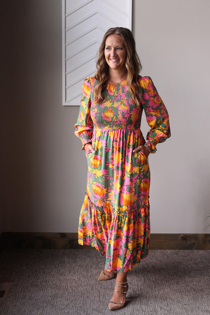 Woman wearing a colorful floral dress standing indoors against a neutral wall.
