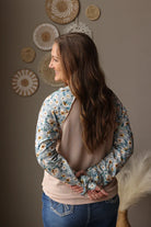 Woman wearing a floral long-sleeve shirt with a neutral sweater underneath, standing against a wall with decorative plates.