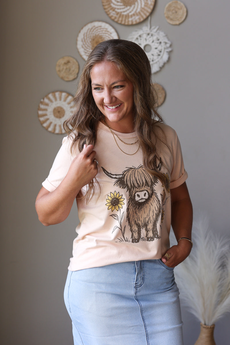 A woman smiling while wearing a light beige graphic t-shirt with a Highland cow and sunflower design, paired with light blue jean skirt, in a styled indoor setting with boho wall decor.