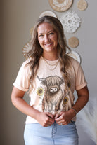 A smiling woman with long light brown hair stands indoors wearing a pale peach graphic T-shirt with a large Highland cow print and blue jean skirt. The background shows a neutral wall with decorative circular wall art.