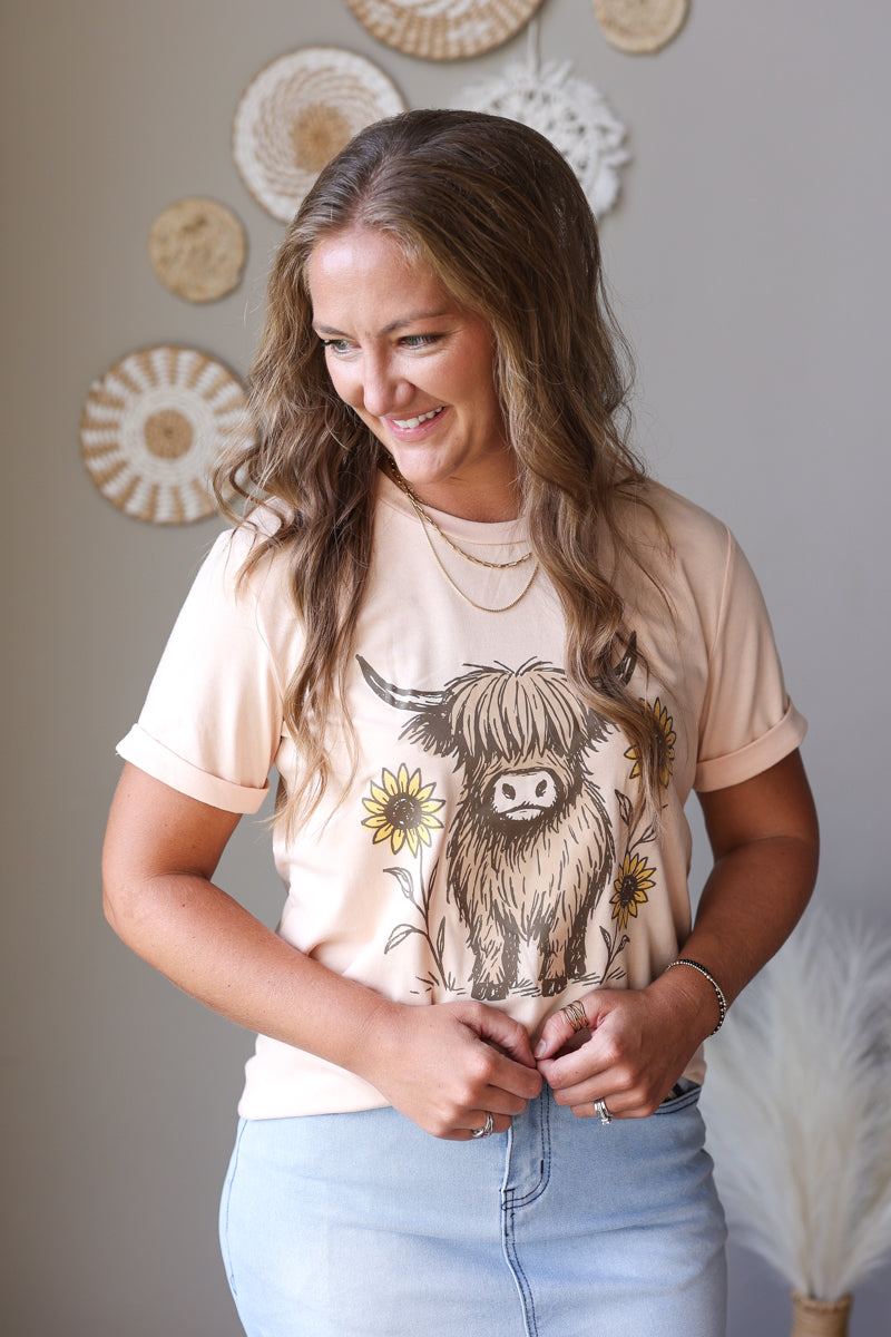 A woman wearing a light peach t-shirt with a graphic of a highland cow and sunflowers, paired with light blue jean skirt, standing in front of a neutral wall with decorative woven wall art.