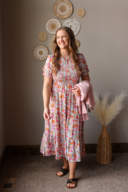 Woman in a floral dress holding a pink jacket in a room with decorative wall art.