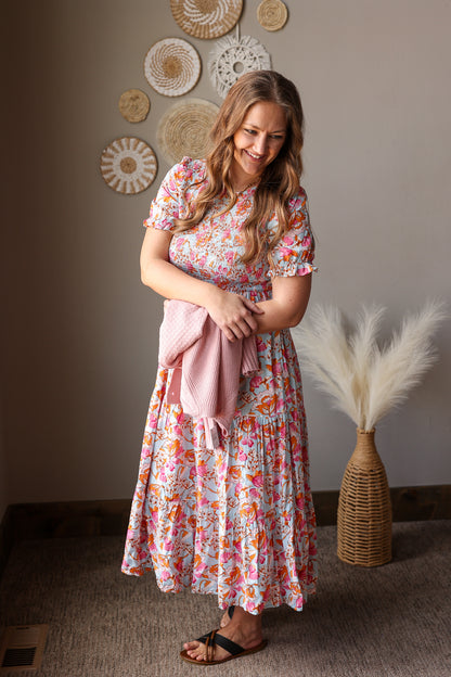 Woman in a floral dress holding a pink towel in a room with decorative wall art and a vase.