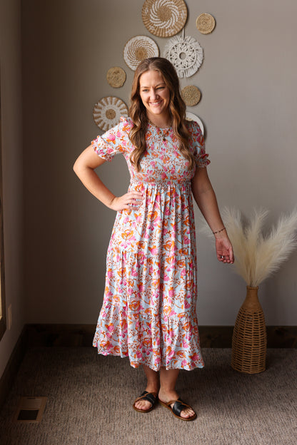 Woman in a floral dress standing in a room with decorative wall art and a vase.
