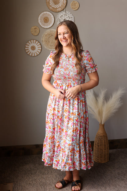 Woman in a floral dress standing in a room with decorative wall art and plants.