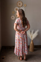 Woman in a floral dress standing in a room with decorative wall art and a vase with dried plants.