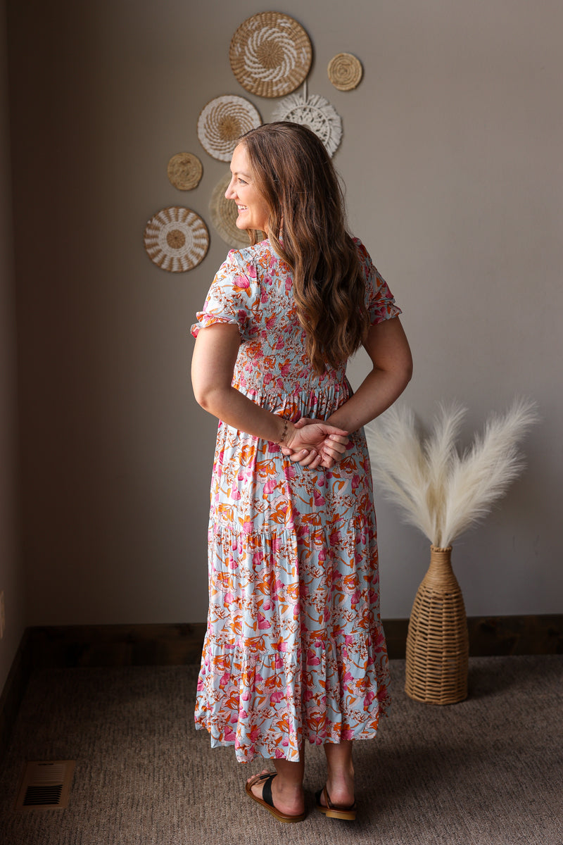 Woman in a floral dress standing in a room with decorative wall art and a vase with dried plants.