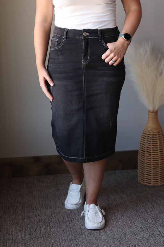Person wearing a black denim skirt with a white top and white sneakers, standing against a neutral background.