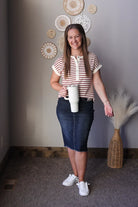 Woman holding a white mug in a room with decorative wall art and a vase, wearing a cream and red striped henley short sleeve top and black wash denim skirt.