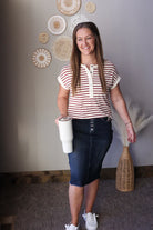 Woman in striped shirt and denim skirt holding a white tumbler in a room with decorative wall art.
