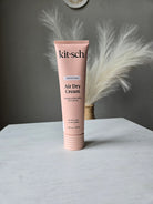 A pink tube of kit·sch Air Dry Cream smoothing air dry shampoo, styled diagonally on a white surface with a white feather prop against a dark gray background.
