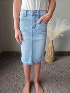 A person wearing a light blue denim mini skirt over a white top, standing on a textured gray carpet with a woven decorative vase and white feather in the background.