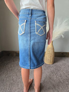 A person wearing a blue denim mini skirt with white embroidered geometric details on the back pockets, standing on a gray carpeted floor.