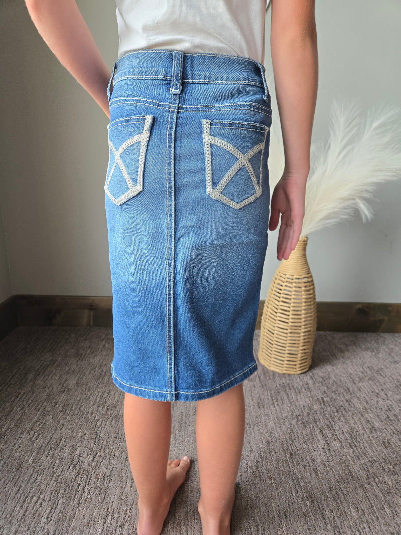 A person wearing a blue denim mini skirt with white embroidered geometric details on the back pockets, standing on a gray carpeted floor.