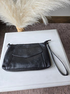 A black leather wristlet clutch with a zippered closure and wrist strap, displayed on a white rectangular box next to a decorative pampas grass stem.