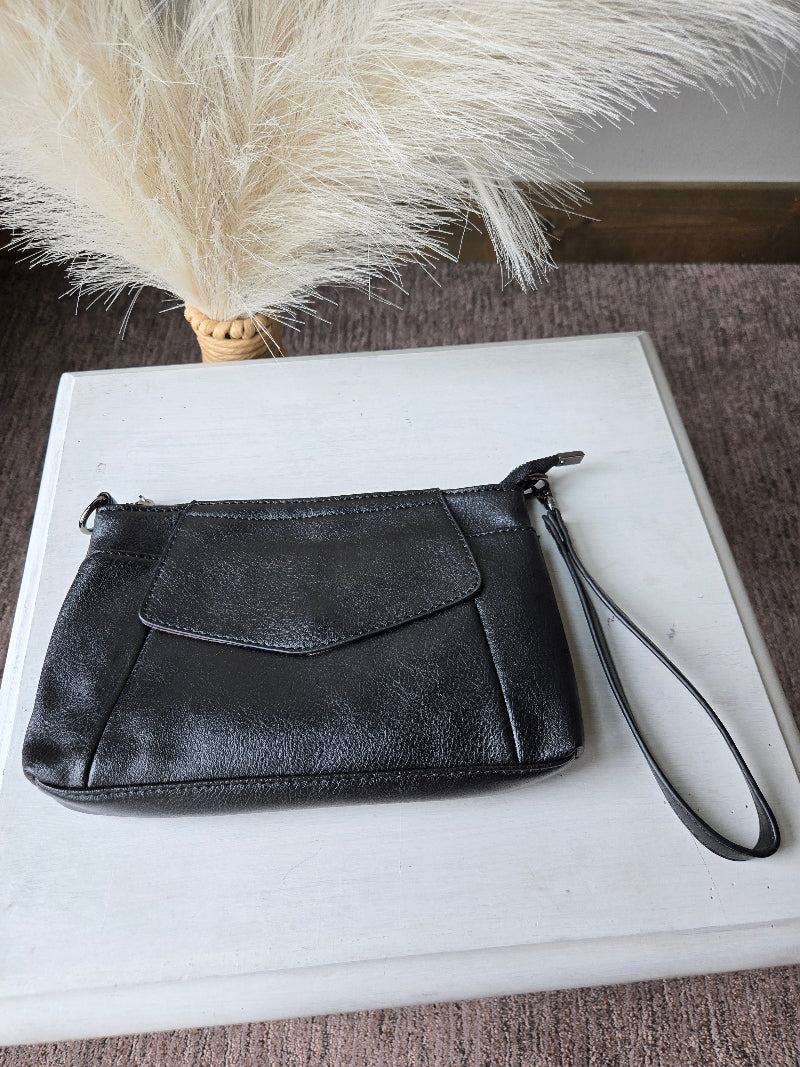 A black leather wristlet clutch with a zippered closure and wrist strap, displayed on a white rectangular box next to a decorative pampas grass stem.