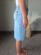 A person wearing light blue denim shorts and a white t-shirt, standing on a gray carpet next to a woven basket and a white feather decoration.