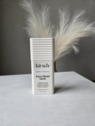 A white rectangular bottle of kit-sch Freshfield Spray with black text, placed on a white surface next to a fluffy white feather against a gray background.
