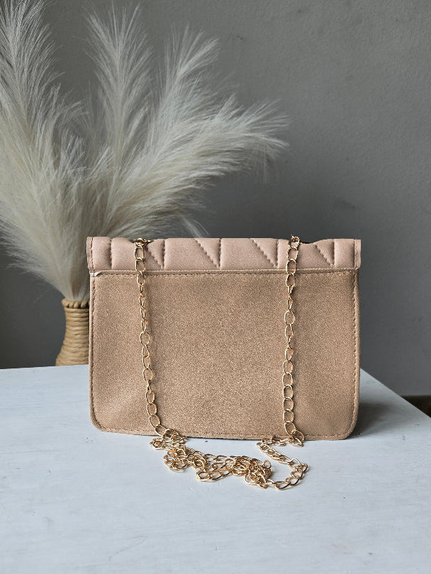 A small structured handbag in a light champagne beige satin-like material, featuring a gold chain strap and decorative gold chain trim, displayed on a white surface next to a white feather prop.