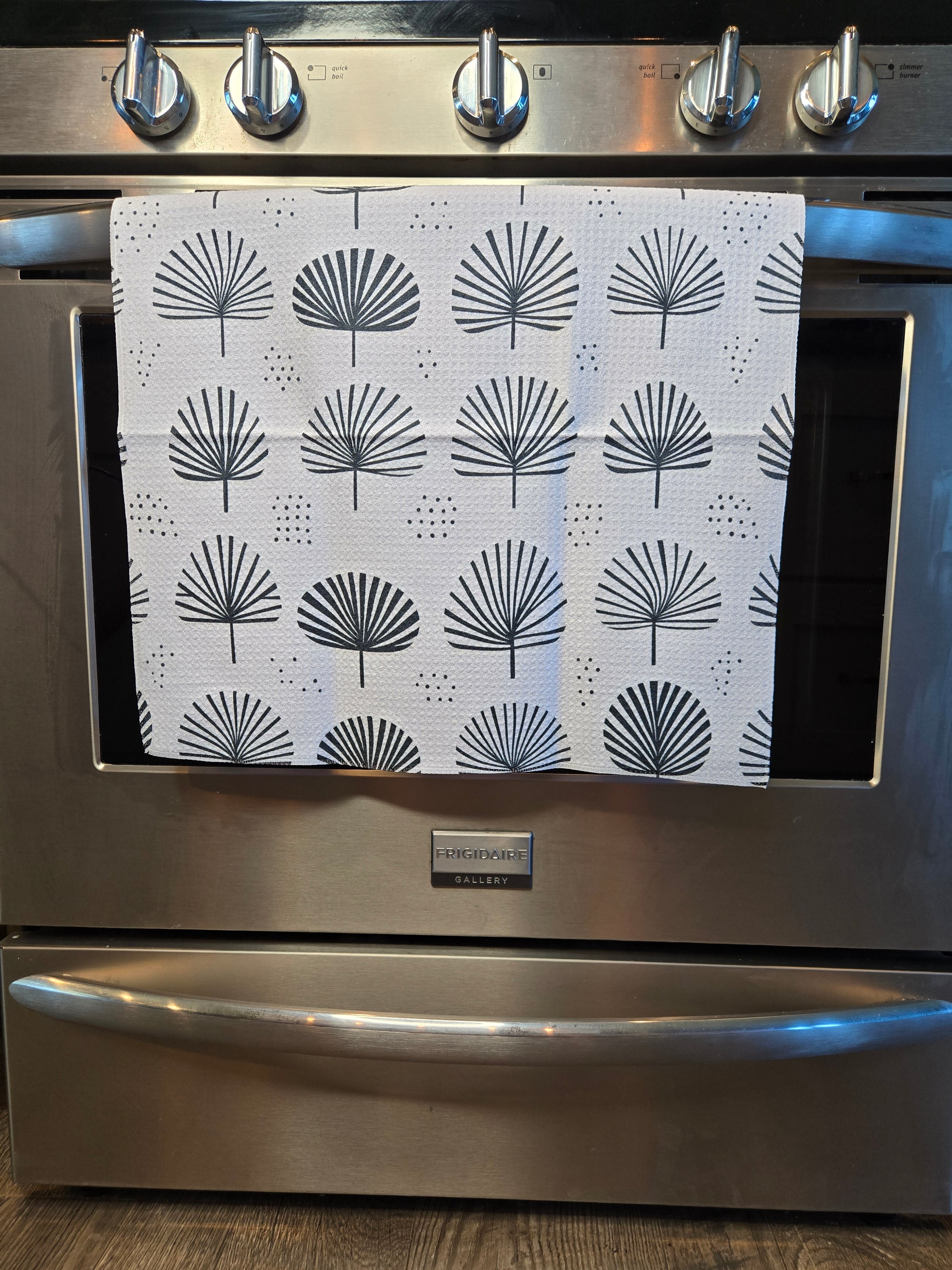 A white kitchen towel with a black palm fan pattern hanging on the oven door of a stainless steel range.